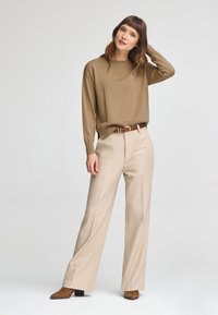 Brauner Pullover mit langen Ärmeln, kombiniert mit beigen weiten Hosen und einem braunen Gürtel. Das Outfit hat ein dezentes Logo auf dem Pullover.