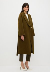 Olivgrön, lång ullrock med breda slag, knäppning med två knappar och sidofickor. Modellen bär figursydda olivfärgade byxor och vita spetsiga klackskor.