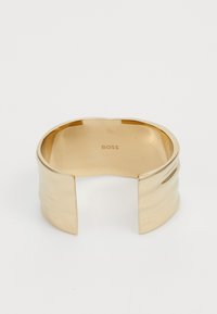 Gullfarget mansjettarmbånd med en glatt, polert overflate. Har bølgende detaljer og en bred åpning. Graverte "BOSS" på innersiden.