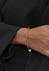 Gouden schakelarmband met een kleine ronde hanger om de pols van een persoon die een zwart langmouwshirt draagt.