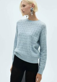 Pulover tricotat de culoare bleu deschis, cu un model texturat, guler rotund, manșete și tiv cu nervuri. Purtați împreună cu o fustă neagră, evidențiind un aspect stratificat.