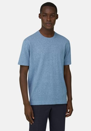 Mand stående med fronten mod kameraet iført en lyseblå kortærmet T-shirt med rund hals og mørke bukser foran en ensfarvet baggrund.