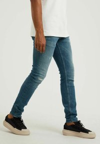 Slim fit modré džíny s distressed detaily, nošeny s černými teniskami s tlustou béžovou podrážkou. Hladká denimová tkanina, ležérní design.