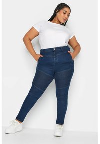 Jeans de color azul oscuro con cintura alta, paneles de rodilla texturizados y corte ceñido. Combinados con una camiseta blanca de manga corta y zapatillas blancas.
