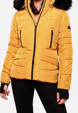 Femme portant une veste d’hiver matelassée jaune moutarde avec une fermeture éclair noire, deux poches avant zippées et une capuche bordée de fourrure noire, associée à un pantalon noir.