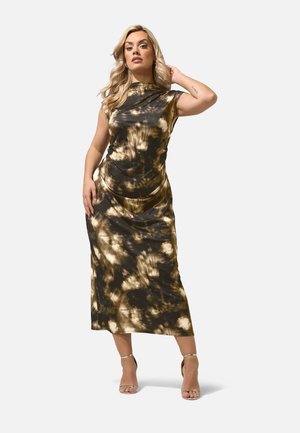 Kvinde med blondt hår iført en ærmeløs, brun og beige tie-dye midi-kjole og gyldne sandaler med remme, poserer med den ene hånd på hovedet.