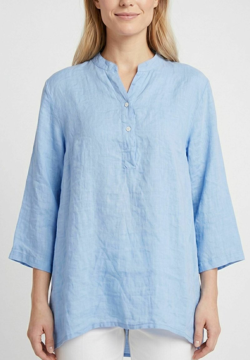 Femme portant une tunique en lin bleu clair à manches longues avec un col mao et deux boutons, associée à un pantalon blanc sur un fond uni.