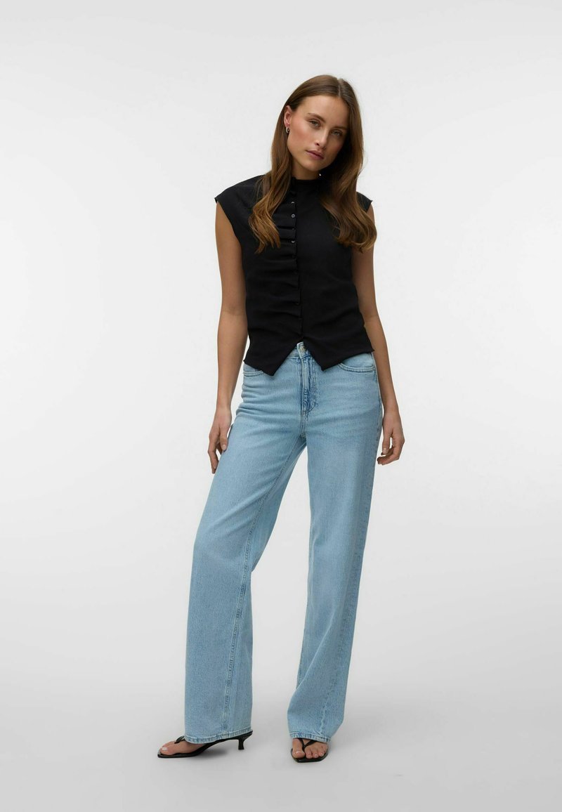 Femme portant un haut noir sans manches boutonné, un jean large taille haute bleu clair et des talons ouverts noirs sur un fond uni.