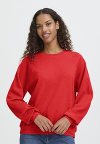 Jongeman met krullend haar die een rode sweatshirt met lange mouwen en kleine kralendetails draagt, staande met de handen zachtjes voor zich gevouwen.