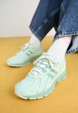 Osoba w jasnozielonych butach do biegania, białych skarpetkach i niebieskich dżinsach siedząca na żółtej powierzchni ze skrzyżowanymi nogami.