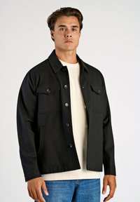 Svart jacka med krage, två bröstfickor och knäppning. Bärs över en ljusfärgad skjorta med en strukturerad tyg.