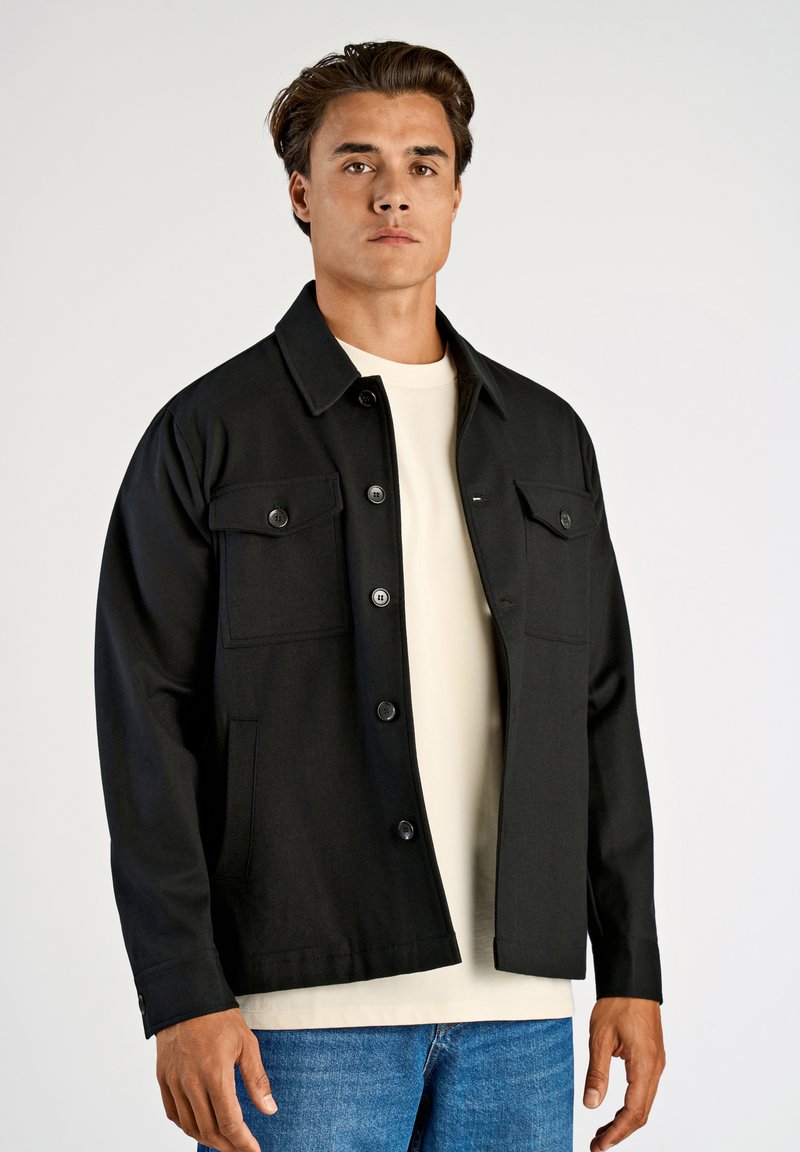 Svart jacka med krage, två bröstfickor och knäppning. Bärs över en ljusfärgad skjorta med en strukturerad tyg.