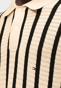 Μπεζ πλεκτό πόλο με μαύρες κάθετες λωρίδες και δύο κουμπιά στη λαιμόκοψη, με ένα μικρό λογότυπο σημαίας σε κόκκινο, άσπρο και μπλε χρώμα.