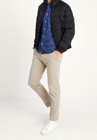 Mężczyzna w czarnej kurtce puchowej, niebieskiej koszuli na guziki, beżowych spodniach i białych trampkach stoi z jedną ręką w kieszeni.