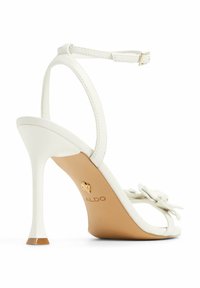 Sandal con tacco alto bianco, con una sottile cinghia alla caviglia, dettaglio floreale sulla punta e una suola beige testurizzata con un logo a forma di cuore.