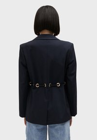 Navy blazer featuring a tailored design, structured shoulders, and metallic eyelet accents at the back, with a smooth texture.