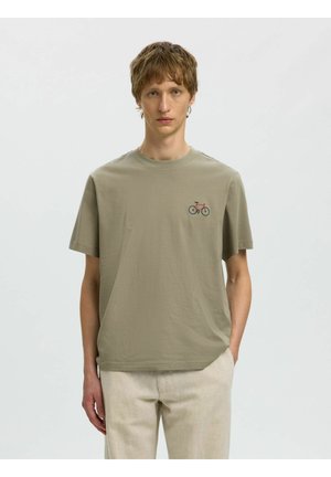 Młody mężczyzna z jasnobrązowymi włosami nosi beżowe spodnie i oliwkowo-zieloną koszulkę z małą czerwoną i niebieską grafiką roweru na piersi.