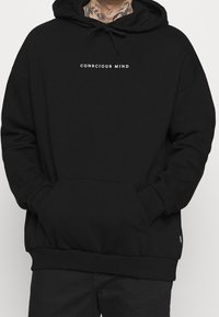 Sudadera negra de mezcla de algodón, con un bolsillo frontal, puños acanalados y capucha con cordón. Texto "MENTE CONSCIENTE" en blanco en el pecho.
