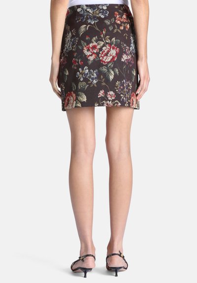 Femme portant une jupe courte foncée avec un motif floral coloré et des sandales noires à talons bas, vue de dos sur fond blanc.