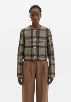 Femme portant une veste à carreaux marron et beige à manches longues boutonnée, avec un pantalon marron uni taille haute, sur un fond uni.