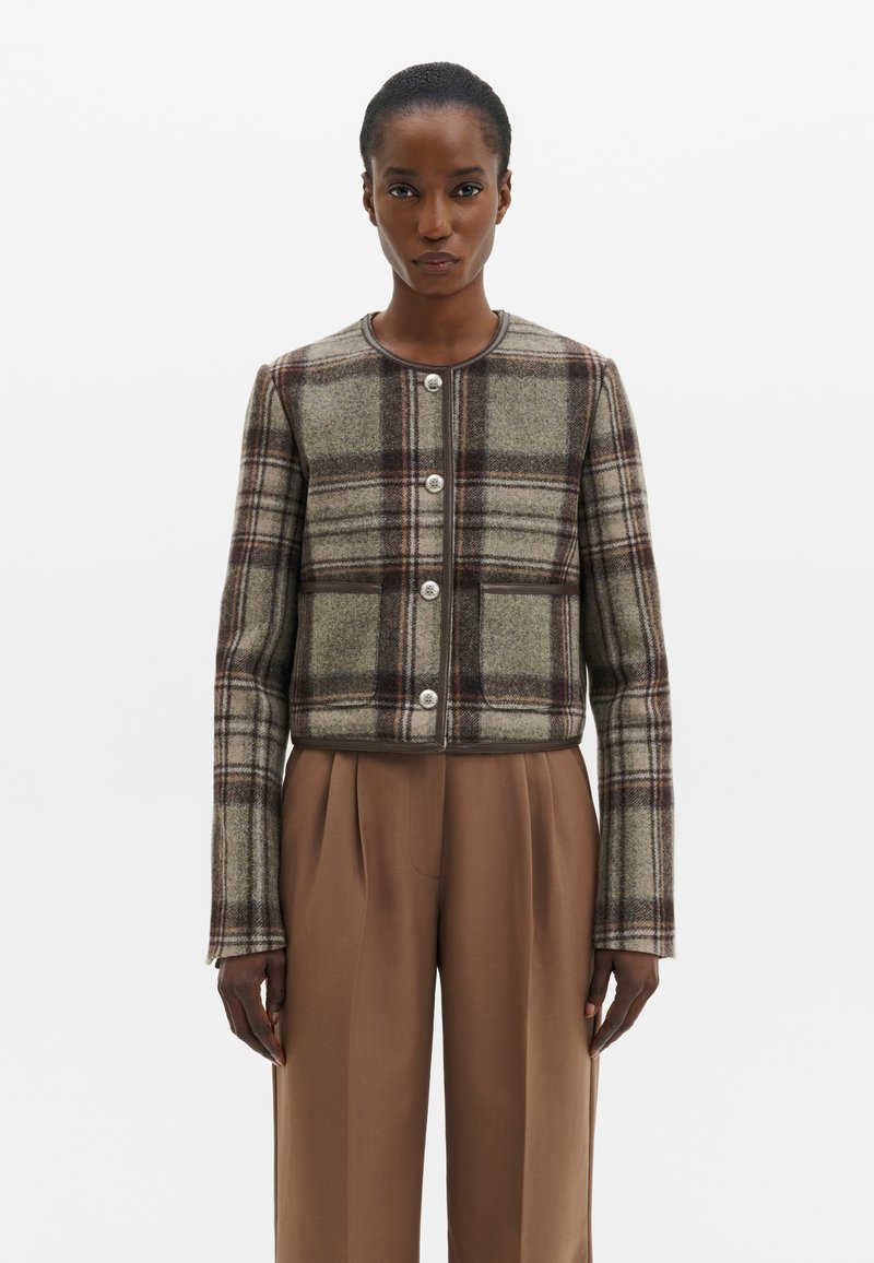 Femme portant une veste à carreaux marron et beige à manches longues boutonnée, avec un pantalon marron uni taille haute, sur un fond uni.