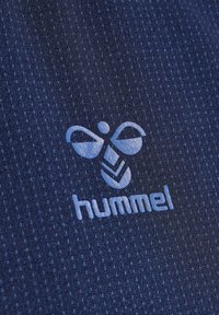 Logo Hummel avec bourdon stylisé au-dessus du nom de la marque en minuscules sur fond de tissu texturé bleu foncé.