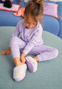 Zapatillas suaves de color pastel y peludas con una suela morada. Un niño se sienta sobre una superficie azul, vistiendo pijamas lavanda con diseños juguetones.
