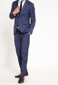 Completo a scacchi blu navy con giacca aderente e pantaloni, caratterizzato da una chiusura con un bottone, abbinato a una camicia bianca e a una cravatta nera. Scarpe in pelle marrone.