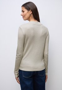 Světlý béžový top s dlouhým rukávem a lesklou texturou, s kulatým límcem a přiléhavým střihem, v kombinaci s modrými džínami.