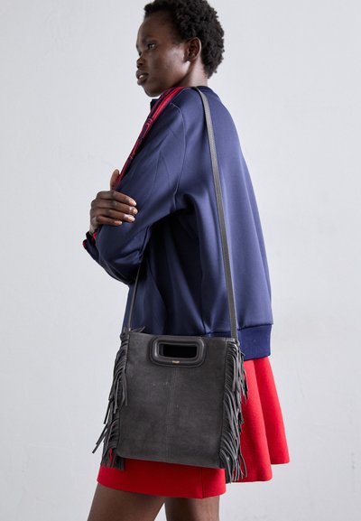 Borsa a tracolla in camoscio grigio con frange, grande apertura con manico superiore e una tracolla sottile. Il modello indossa una giacca blu navy e una gonna rossa.