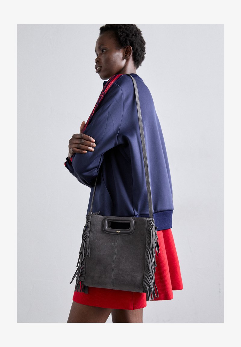 Szara torebka z zamszu z frędzlami, duży otwór z górnym uchwytem i cienki pasek. Modelka nosi granatową kurtkę i czerwoną spódnicę.