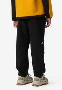 Crne trenirka ima sužene nogavice i uvezeni logo. Model nosi bež planinarske cipele s teksturiranim dizajnom. Iznad je žuta flis jakna.