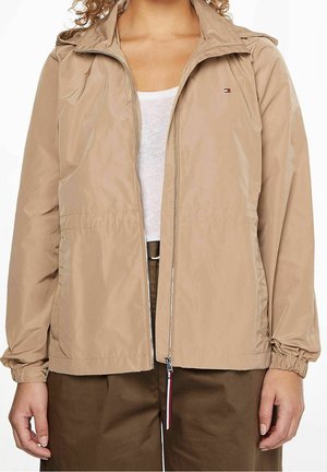 Personne portant une veste beige zippée sur une chemise blanche et un pantalon marron, avec des poignets élastiques et un petit logo sur la poitrine.
