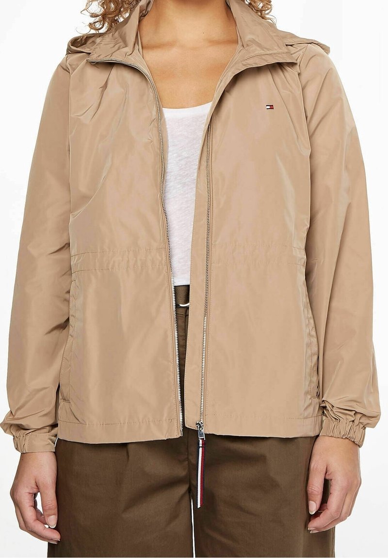 Personne portant une veste beige zippée sur une chemise blanche et un pantalon marron, avec des poignets élastiques et un petit logo sur la poitrine.