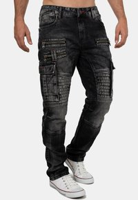 Black denim cargo pants with multiple zippered pockets, ribbed accents on thighs, and contrasting stitch details. Worn with white sneakers.