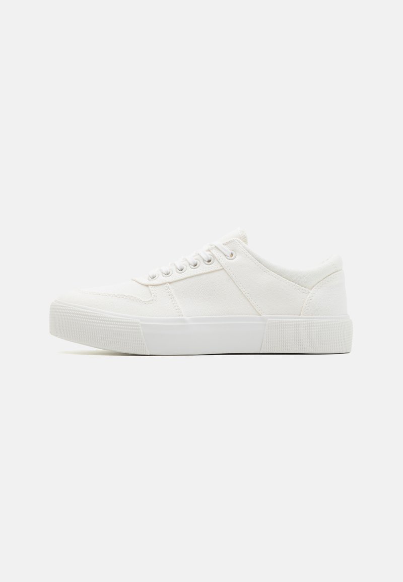 White canvas sneakers with a textured rubber sole, featuring a round toe, lace-up design, and minimalistic accents.