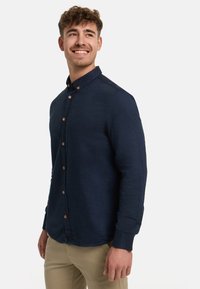 Lächelnder junger Mann mit kurzen lockigen Haaren, der ein marineblaues Hemd mit Knöpfen und langen Ärmeln sowie beige Hosen trägt und zur Seite schaut.