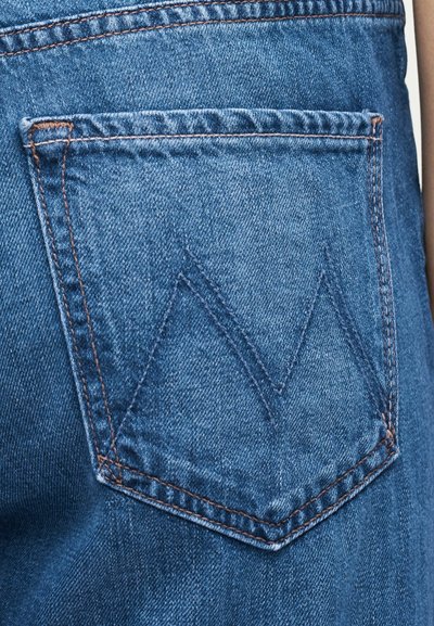 Κοντινή λήψη πίσω τσέπης τζιν από μπλε ντένιμ με διπλή ραφή σε σχήμα "M" και πορτοκαλοκαφέ κλωστή στο ισχίο ενός ατόμου.