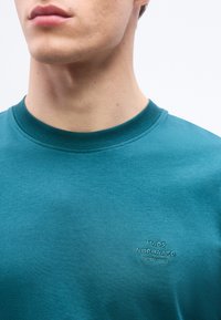 Κοντινό πλάνο ενός ατόμου που φοράει μια μπλουζα με στρογγυλή  λαιμόκοψη σε τιρκουάζ χρώμα, με την κεντημένη φράση "MADS NØRGAARD COPENHAGEN" στο στήθος.