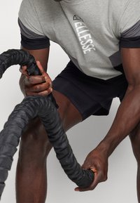 Grå och svart atletisk t-shirt med logotyp och geometrisk design, tillsammans med svarta shorts. Personen håller ett tjockt svart stridsrep.