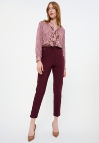 Blouse à motif rouge avec un col noué, associée à un pantalon bordeaux taille haute et des escarpins nude, créant une silhouette épurée.