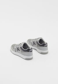 Pár šedých a bílých tenisek New Balance s černým logem na boku a značkou na patě, zobrazený ze zadního úhlu.