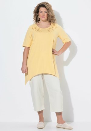 Frau mit lockigem Haar, die eine gelbe Tunika mit floralen Ausschnitten, weiße Caprihose und leichte Slipper trägt, mit einer Hand auf der Hüfte stehend.