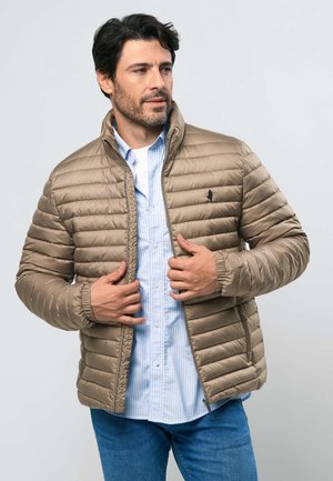 Mand iført beige quiltet jakke over lyseblå stribet skjorte og blå jeans, justerer jakke lynlås, ser til siden.