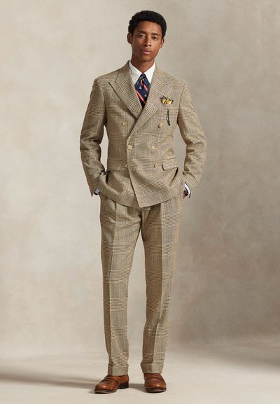 Uomo in piedi con le mani in tasca, indossa un abito doppiopetto a quadri beige, cravatta blu navy con motivi, camicia bianca, scarpe marroni e un fazzoletto da taschino.