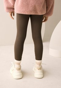 Leggings marroni a coste, scarpe da ginnastica bianche con accenti beige e giacca in pile rosa. Lo sfondo presenta trame e forme morbide.