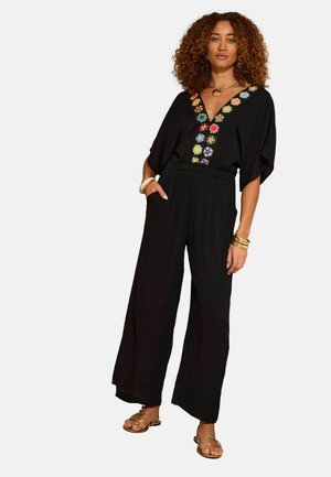 Žena kučeravými vlasmi oblečená v čiernom overal s širokými nohavicami a farebnou kvetinovou výšivkou na V-výstrihu, doplnená zlatými šperkami a sandálmi.