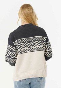 Persona bionda che indossa un maglione lavorato a maglia con un motivo in bianco e nero e design geometrici, abbinato a jeans blu, mostrata di spalle.