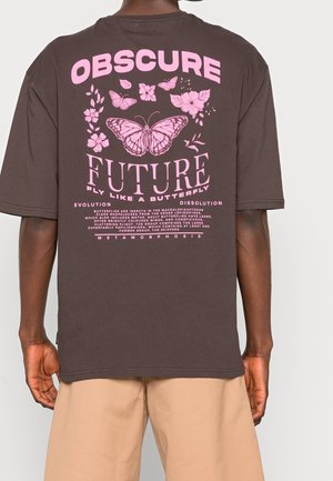 Henkilö, joka pukeutuu tummanruskeaan T-paitaan, jossa on vaaleanpunaisia perhonen ja kukka -grafiikoita sekä teksti "Hämärä Tulevaisuus, Lentää Kuten Perhonen."