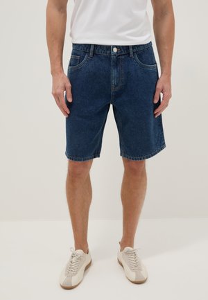 Uomo che indossa pantaloncini di jeans blu scuro, scarpe da ginnastica bianche e una maglietta bianca, in piedi contro uno sfondo chiaro e semplice.